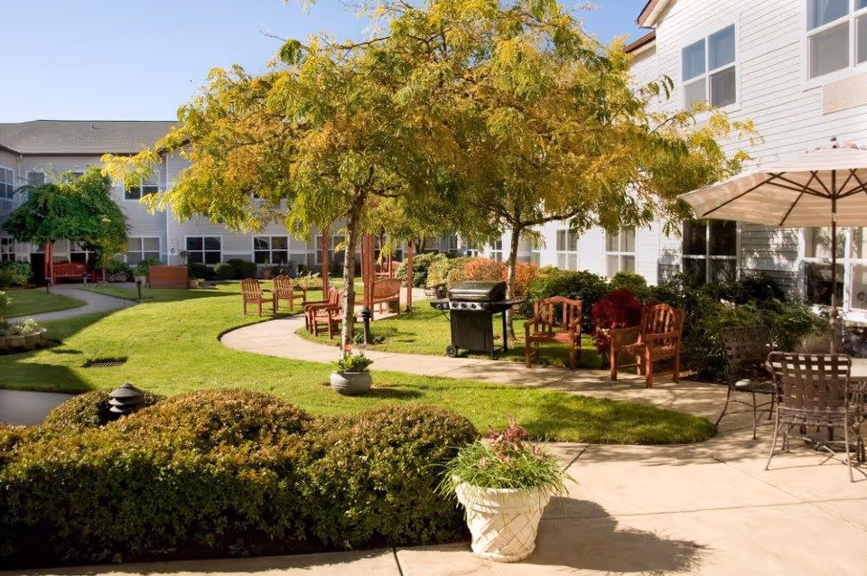Outdoor courtyard area at Merrill Gardens at Sheldon Park featuring green grass, trees, bushes, wooden chairs, a barbecue grill, and a patio table with an umbrella, surrounded by a two-story building.