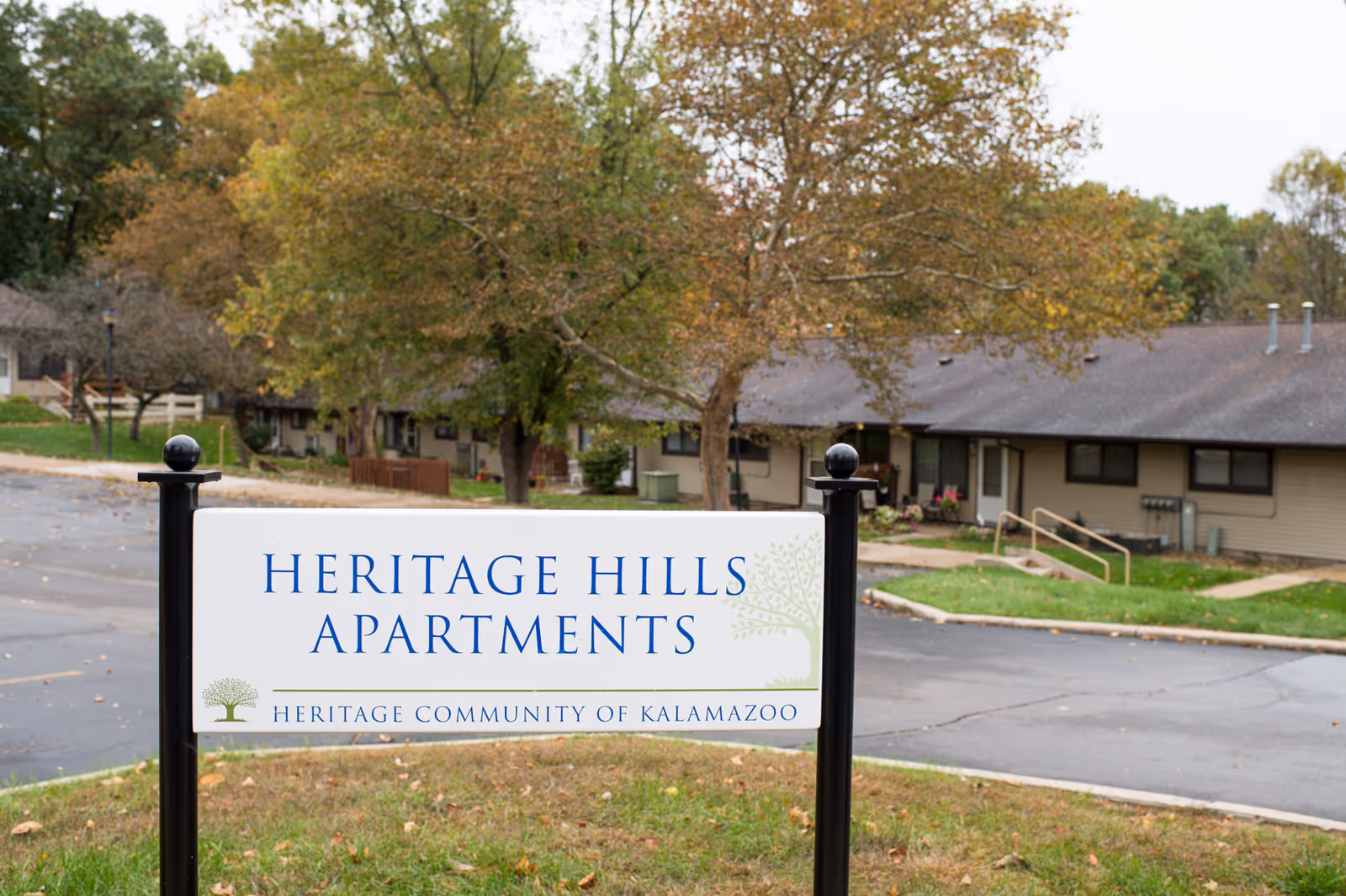 A sign reading 'Heritage Hills Apartments, Heritage Community of Kalamazoo' is displayed in front of a residential apartment complex with trees and a paved driveway in the background.