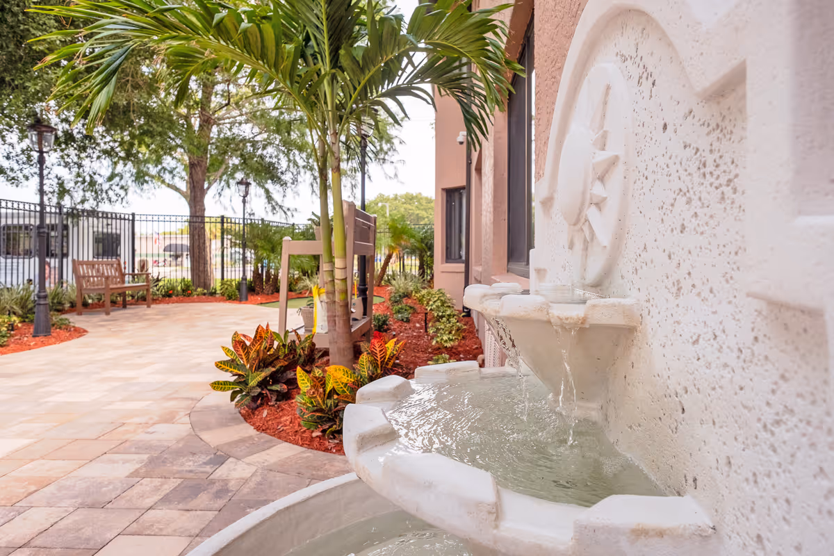 Outdoor garden area with a multi-tiered stone water fountain in the foreground, tropical plants, palm trees, benches, and a paved walkway surrounded by a black metal fence.