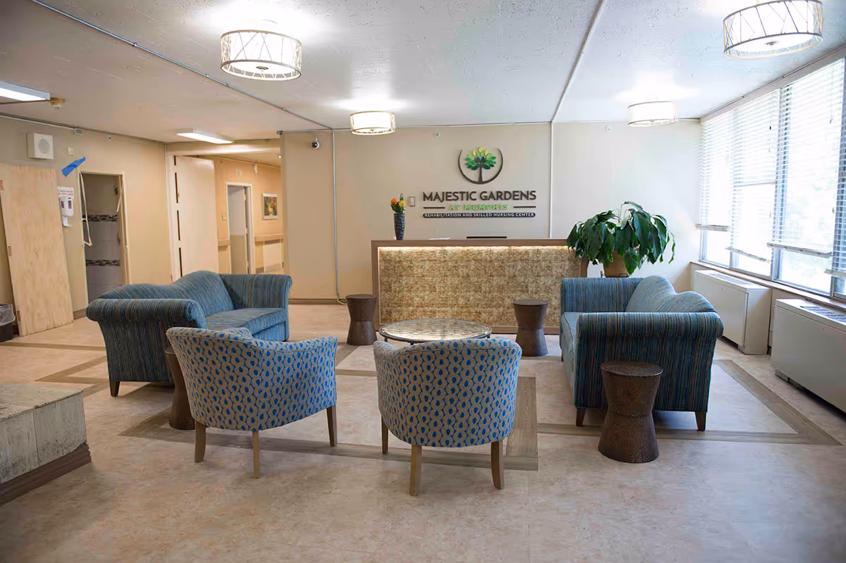 A bright and clean reception area at Majestic Gardens at Memphis Rehabilitation and Skilled Nursing Center featuring a seating arrangement with two blue patterned armchairs, two blue striped loveseats, a round glass coffee table, and small wooden side tables. The reception desk is in the background with a plant on one side and a vase with flowers on the other. Large windows on the right side allow natural light to fill the room.