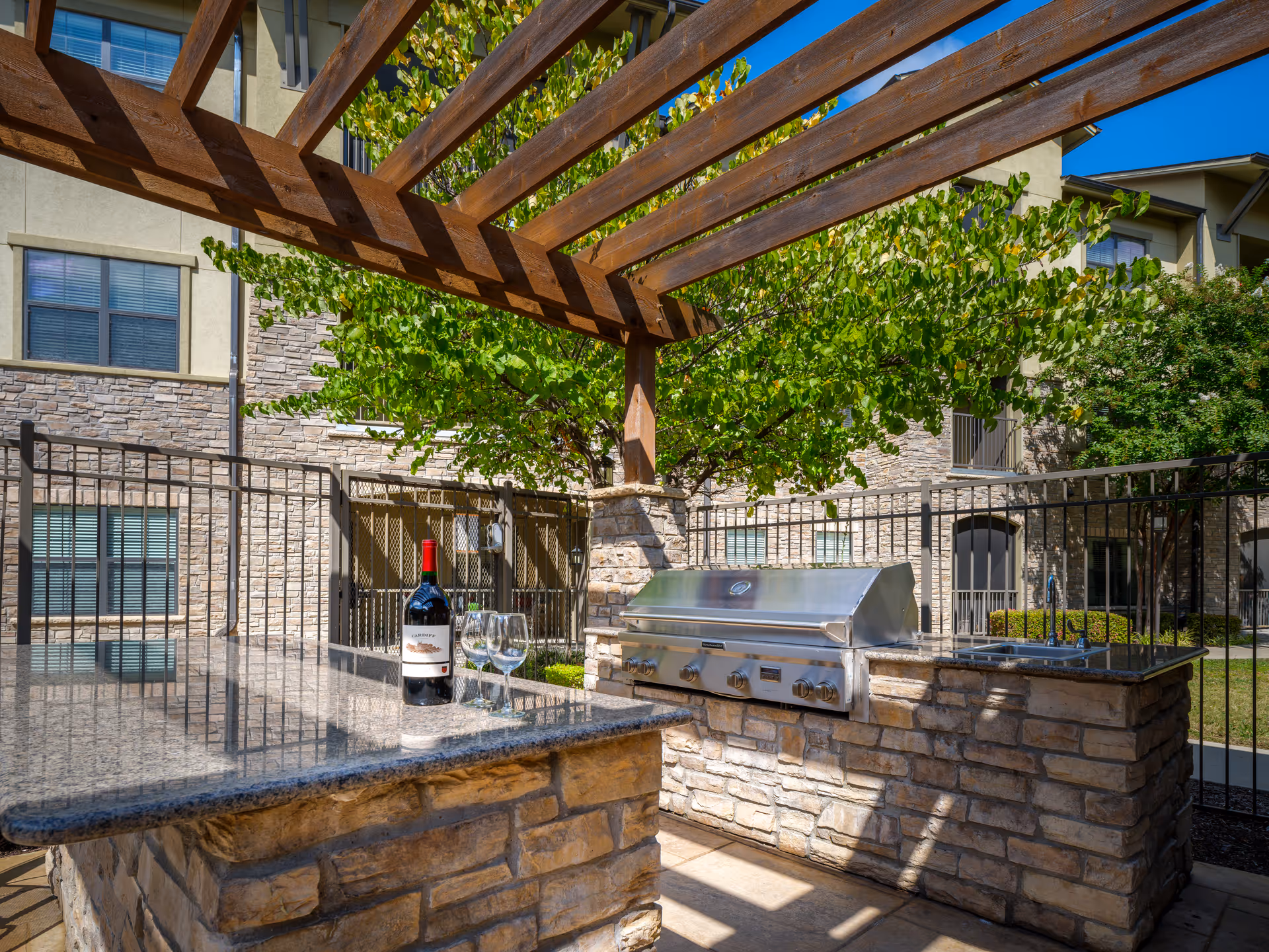 Outdoor patio area with a stone countertop and built-in stainless steel grill under a wooden pergola. A bottle of red wine and two wine glasses are placed on the countertop. The background shows a fenced area with trees and a multi-story building with stone and beige walls.