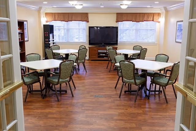 A well-lit common room with several square tables, each surrounded by four green cushioned chairs. The room has wooden flooring, two large windows with valances, and a large flat-screen TV on a wooden stand against the far wall.