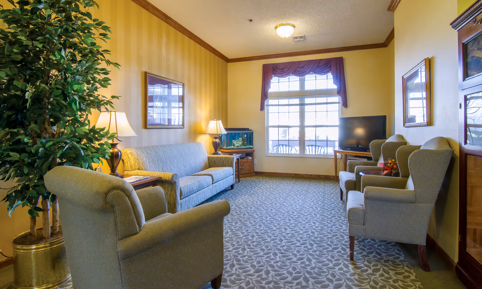 A cozy living room with patterned carpet, a large window with burgundy valance, a blue sofa, three green armchairs, two table lamps, a fish tank on a wooden stand, a flat-screen TV on a wooden table, framed artwork on the walls, and a large potted plant in a brass container.