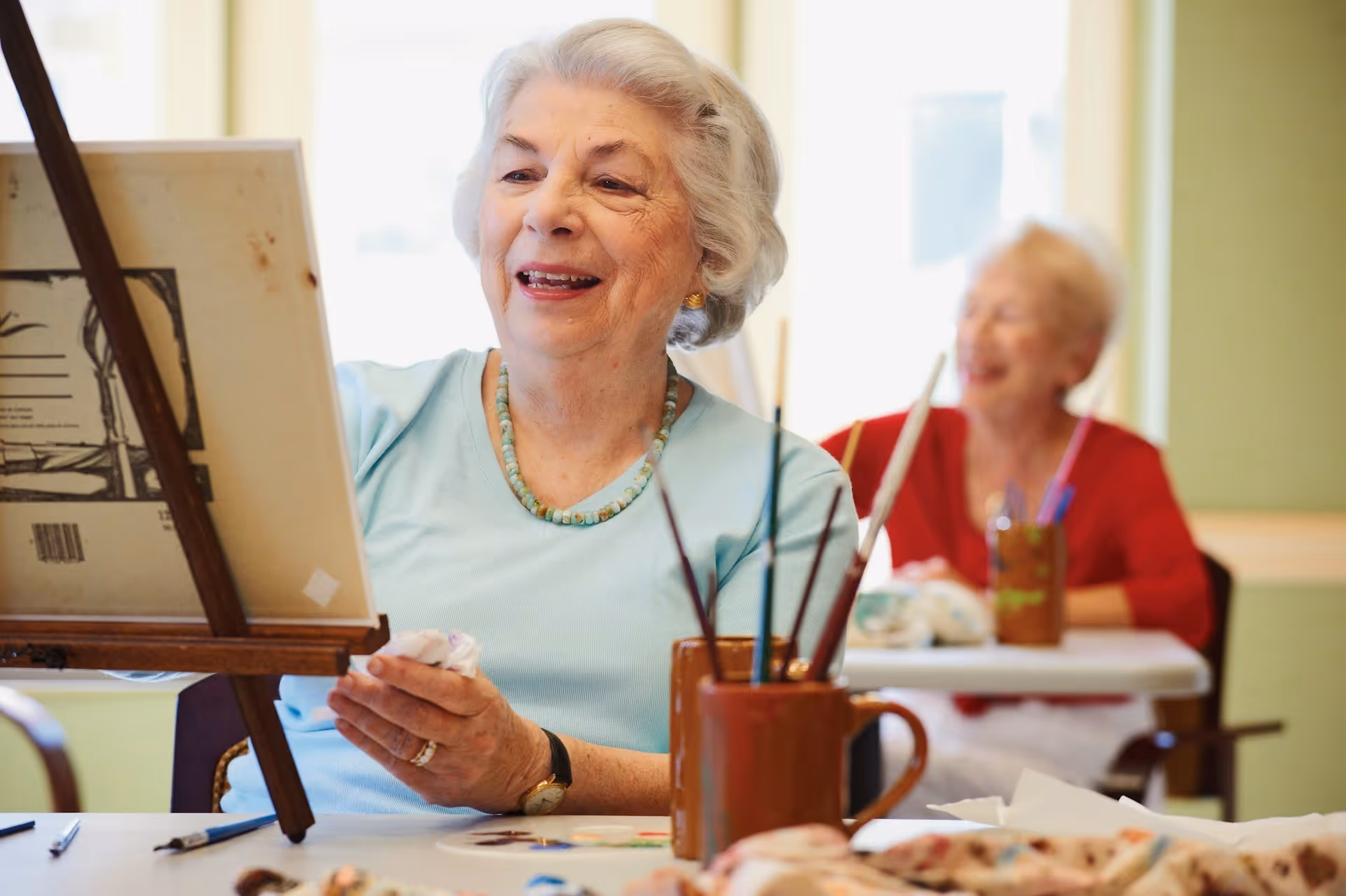 Two elderly women engaged in painting activities at a table inside a well-lit room. The woman in the foreground is smiling and holding a paintbrush, working on a canvas on an easel. The woman in the background is also smiling and appears to be enjoying the activity.