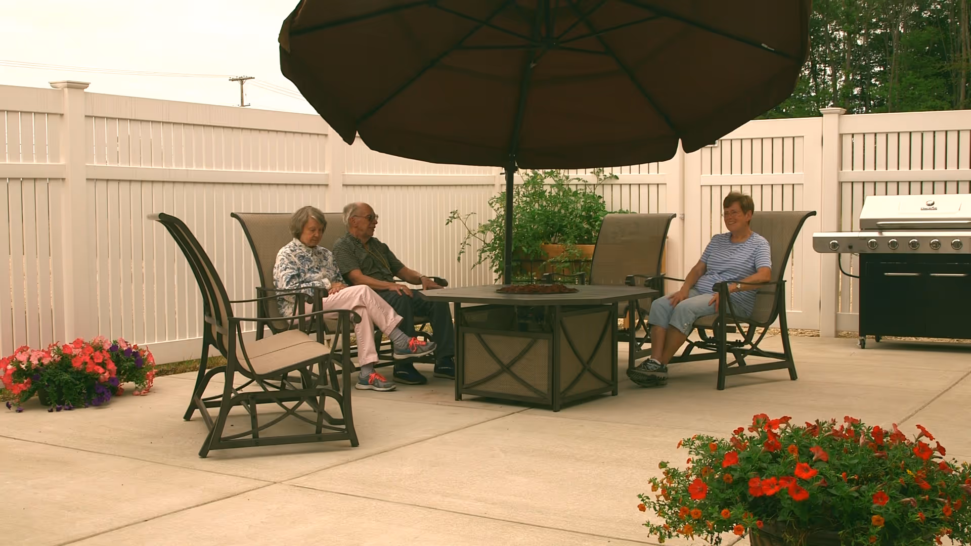 Three elderly people sitting on patio chairs around a round table with a large umbrella in an outdoor area enclosed by a white fence. There are flower pots with colorful flowers and a barbecue grill in the background.