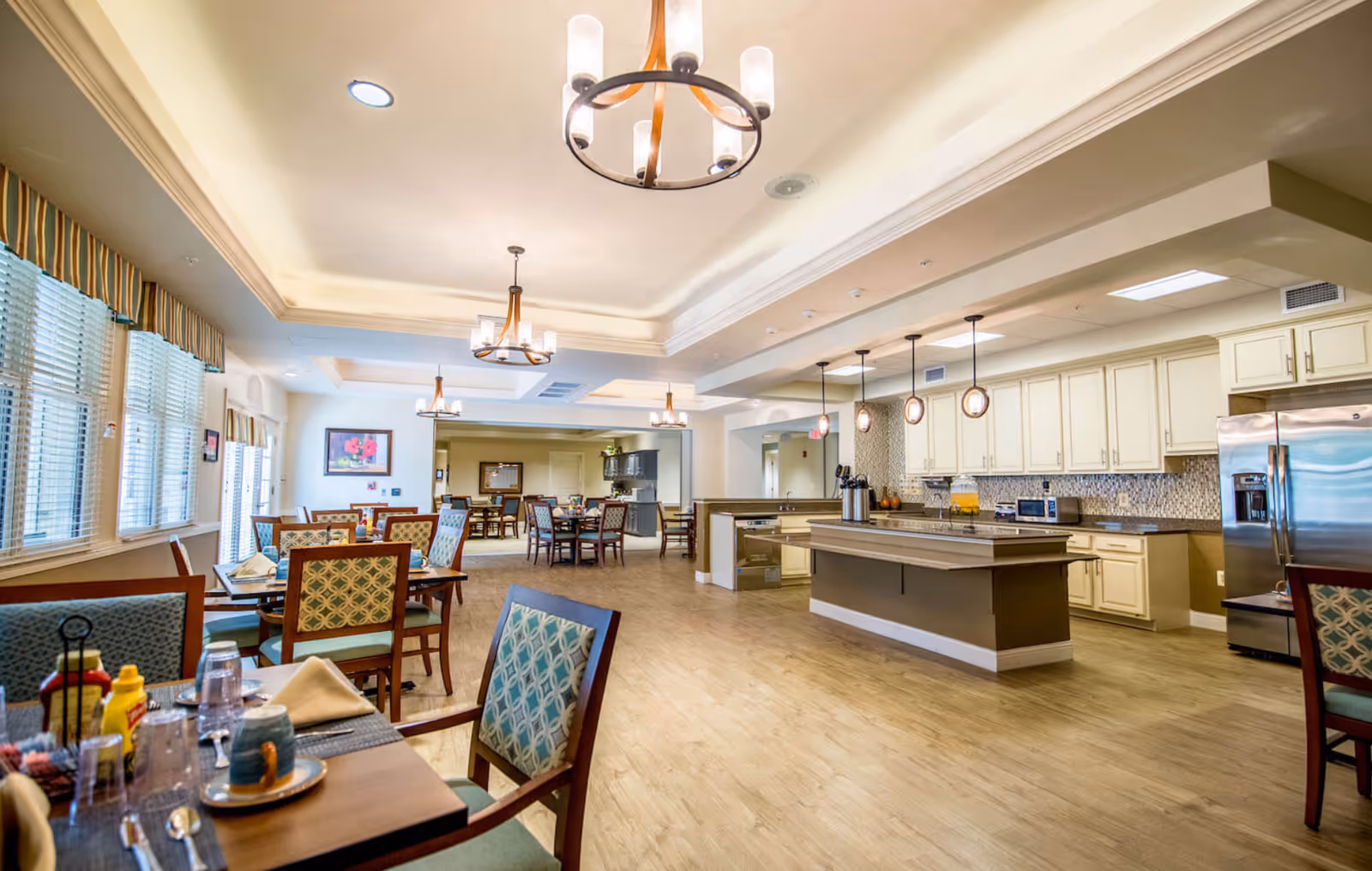 A spacious and well-lit dining area in Chantilly Heights Memory Care featuring multiple wooden tables and chairs with patterned upholstery. The room has large windows with blinds and valances, modern chandeliers hanging from a tray ceiling, and a kitchen area with cream-colored cabinets, stainless steel refrigerator, and a central island with pendant lights.