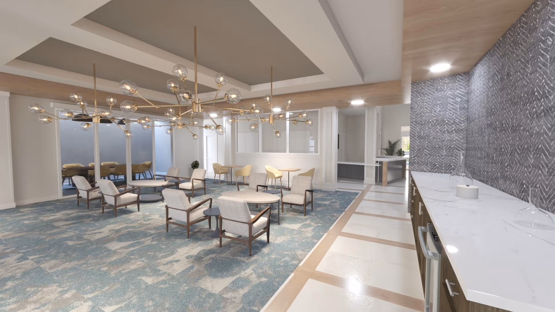 A modern common area with multiple seating arrangements including cushioned armchairs around circular tables on a blue patterned carpet. The ceiling features two large, decorative chandeliers with multiple glass bulbs. To the right, there is a white marble countertop with a gray patterned wall behind it and wooden cabinetry below. The floor near the countertop is tiled with a light color and wooden accents. Large windows and doors are visible in the background, allowing natural light into the space.