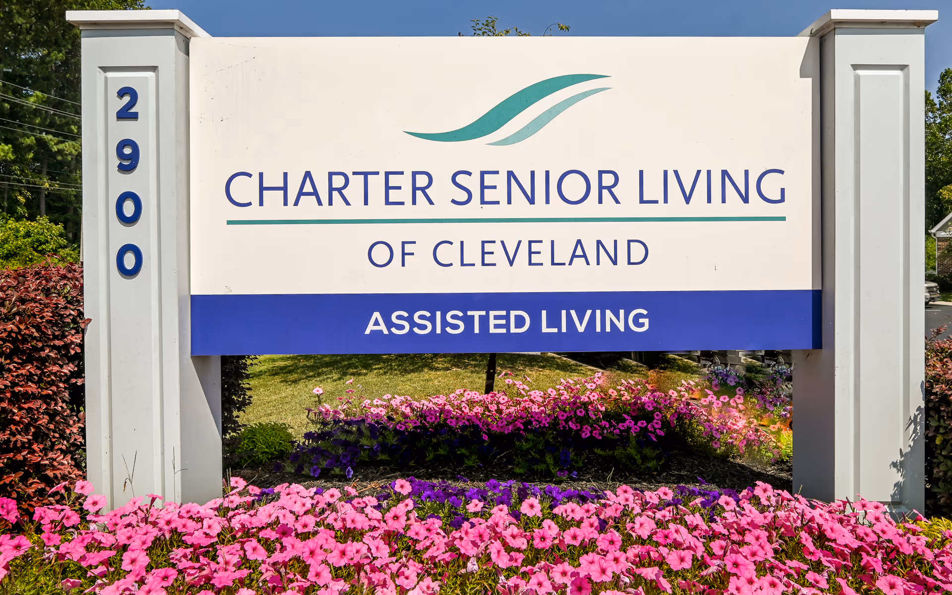Outdoor sign for Charter Senior Living of Cleveland Assisted Living, surrounded by vibrant pink and purple flowers with green foliage and a clear blue sky in the background.