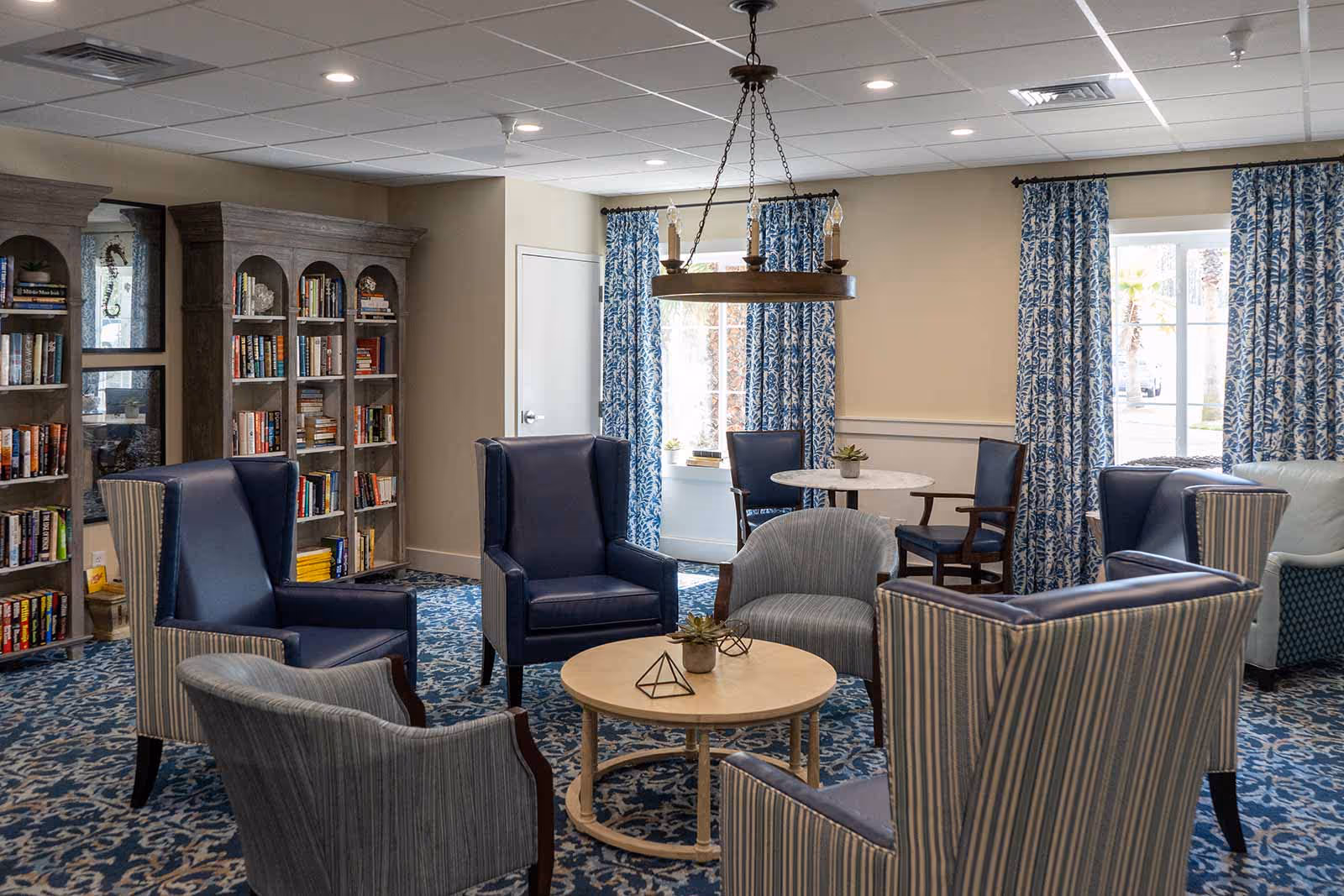 A cozy sitting area in a senior living facility with several upholstered armchairs arranged around a round wooden coffee table. Behind the seating area, there are tall bookshelves filled with books and decorative items. The room has blue patterned carpet and matching blue patterned curtains on the windows, with a round table and chairs near the windows. A chandelier hangs from the ceiling providing light.