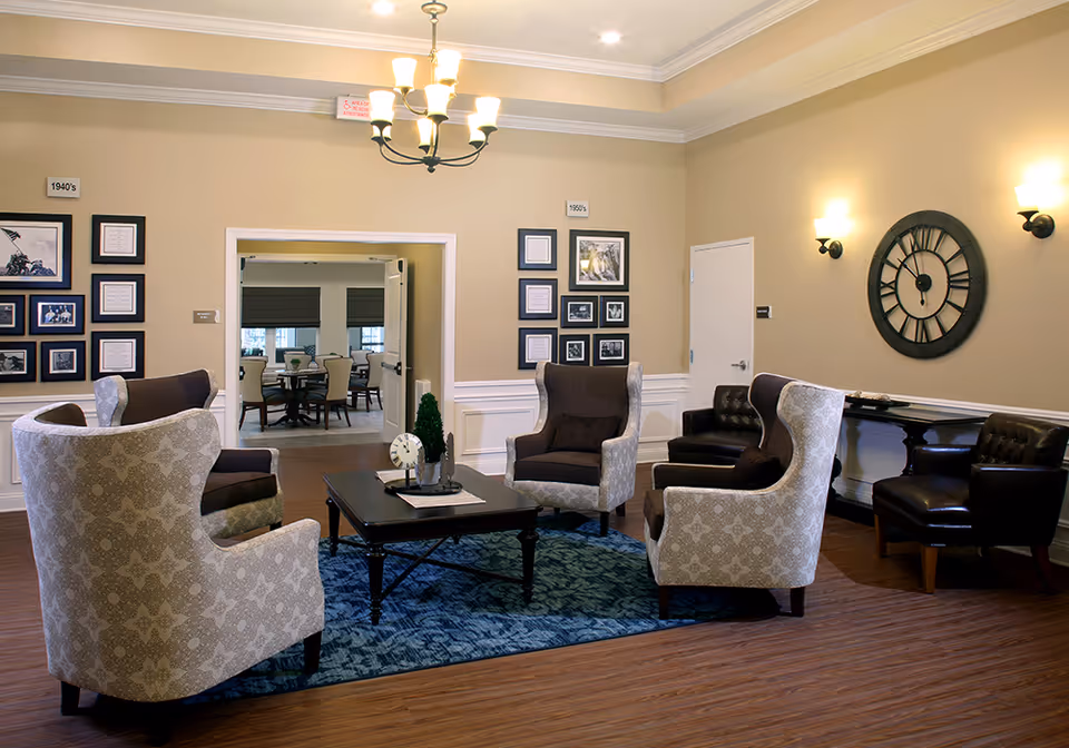 A cozy living room area with four patterned armchairs arranged around a dark wooden coffee table on a blue rug. The walls are beige with white wainscoting, decorated with framed photos and documents labeled 1940's and 1950's. A large black wall clock and two wall sconces are visible on the right wall. In the background, an adjoining room with a round table and chairs can be seen.