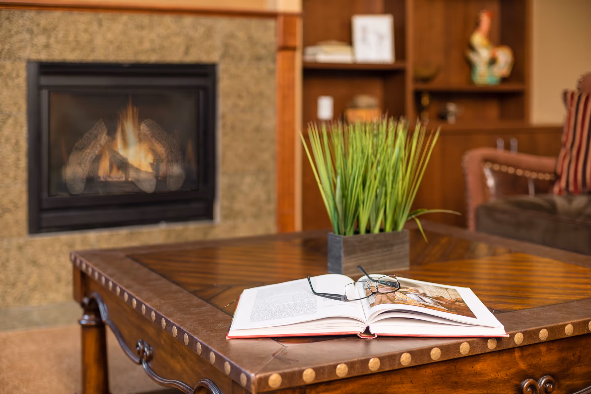 A cozy living room scene featuring a wooden coffee table with an open book and a pair of reading glasses resting on it. A rectangular planter with green grass-like plants is also on the table. In the background, there is a lit fireplace with a stone surround and wooden shelving with decorative items, including a rooster figurine and framed pictures. A cushioned armchair with striped upholstery is partially visible.