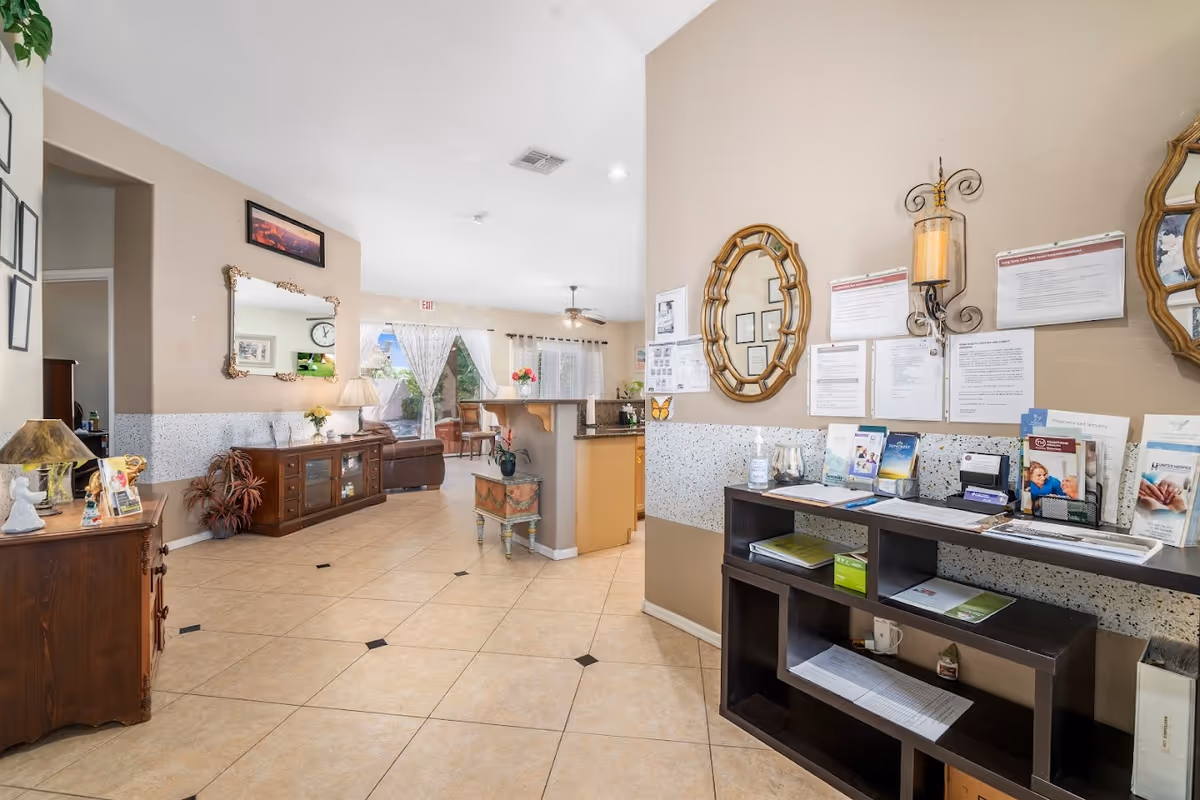 Interior view of a senior living facility lobby or common area with tiled floors, beige walls, and various wooden furniture including cabinets and shelves. There are informational brochures and notices on the wall, a decorative mirror, and a small table with a vase of flowers. In the background, there is a seating area with chairs and large windows with curtains letting in natural light.