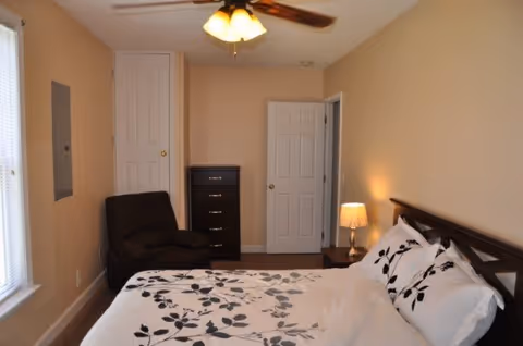 A bedroom with a bed featuring white bedding with a black leaf pattern, a dark wooden headboard, a bedside table with a lamp, a dark armchair, a tall chest of drawers, and two closed white doors. The walls are beige and there is a ceiling fan with a light fixture.