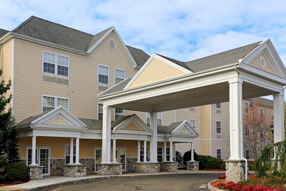 Covered front entrance of a multi-story beige senior living building with white columns and stone bases under a porte-cochère.