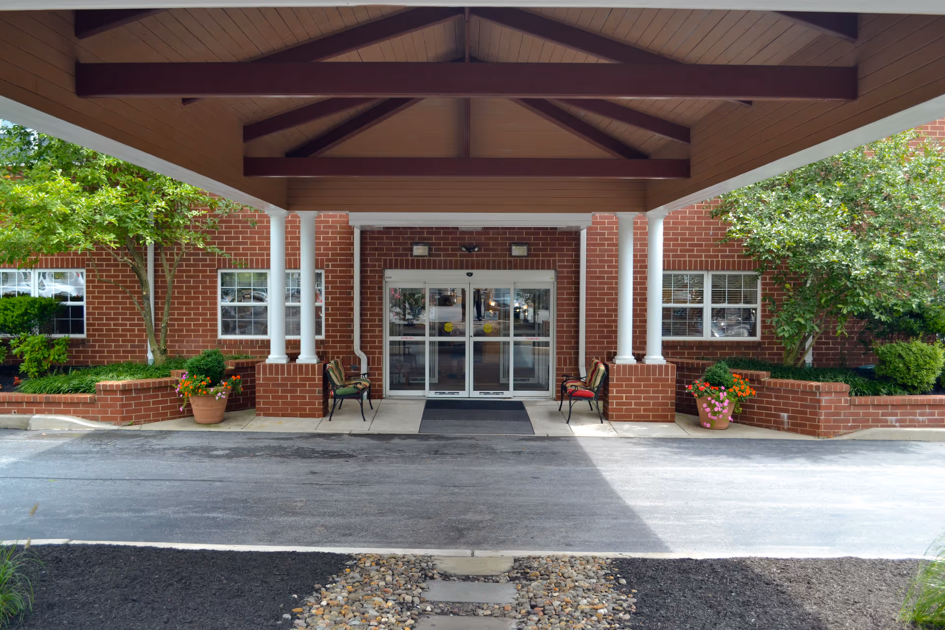 Covered entrance to a brick building with glass double doors, two chairs on either side, potted plants, and greenery around the entrance.