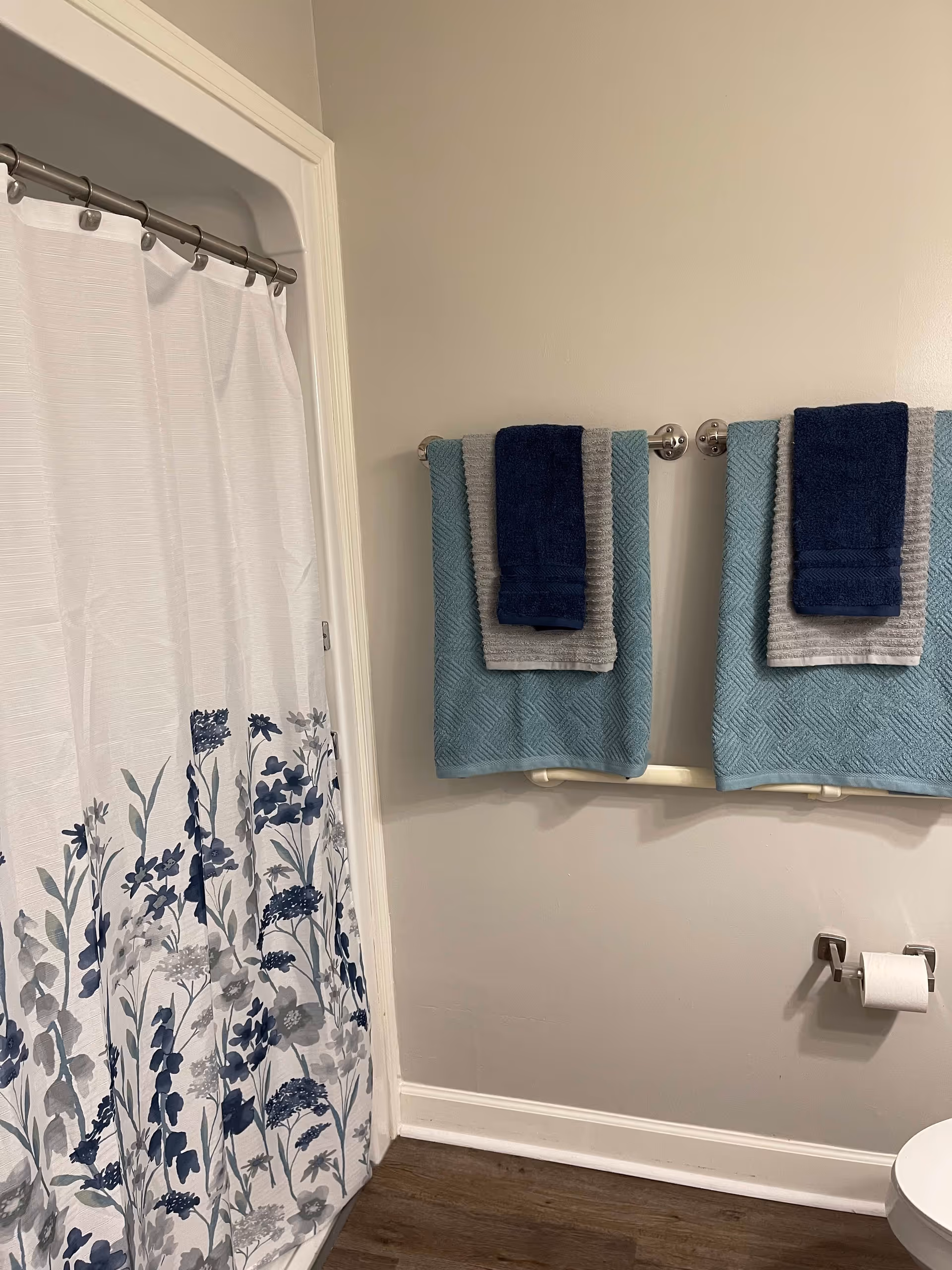 Bathroom interior with a white shower curtain featuring blue and gray floral patterns, two towel racks each holding a blue, gray, and navy towel, a toilet paper holder with a roll of toilet paper, and wood-look flooring.