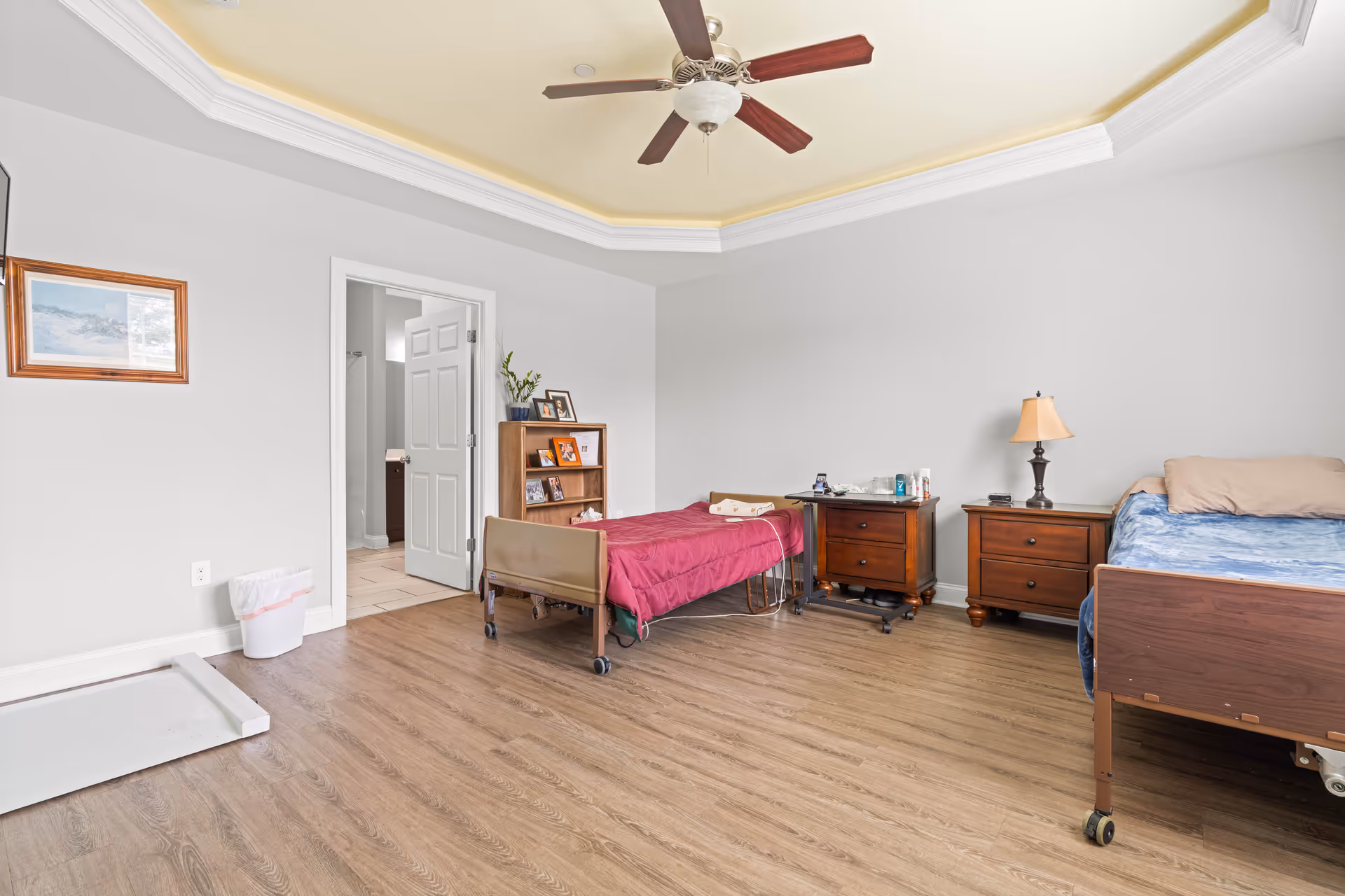 Spacious assisted-living bedroom with two beds, nightstands, a ceiling fan, and a doorway leading to a bathroom.