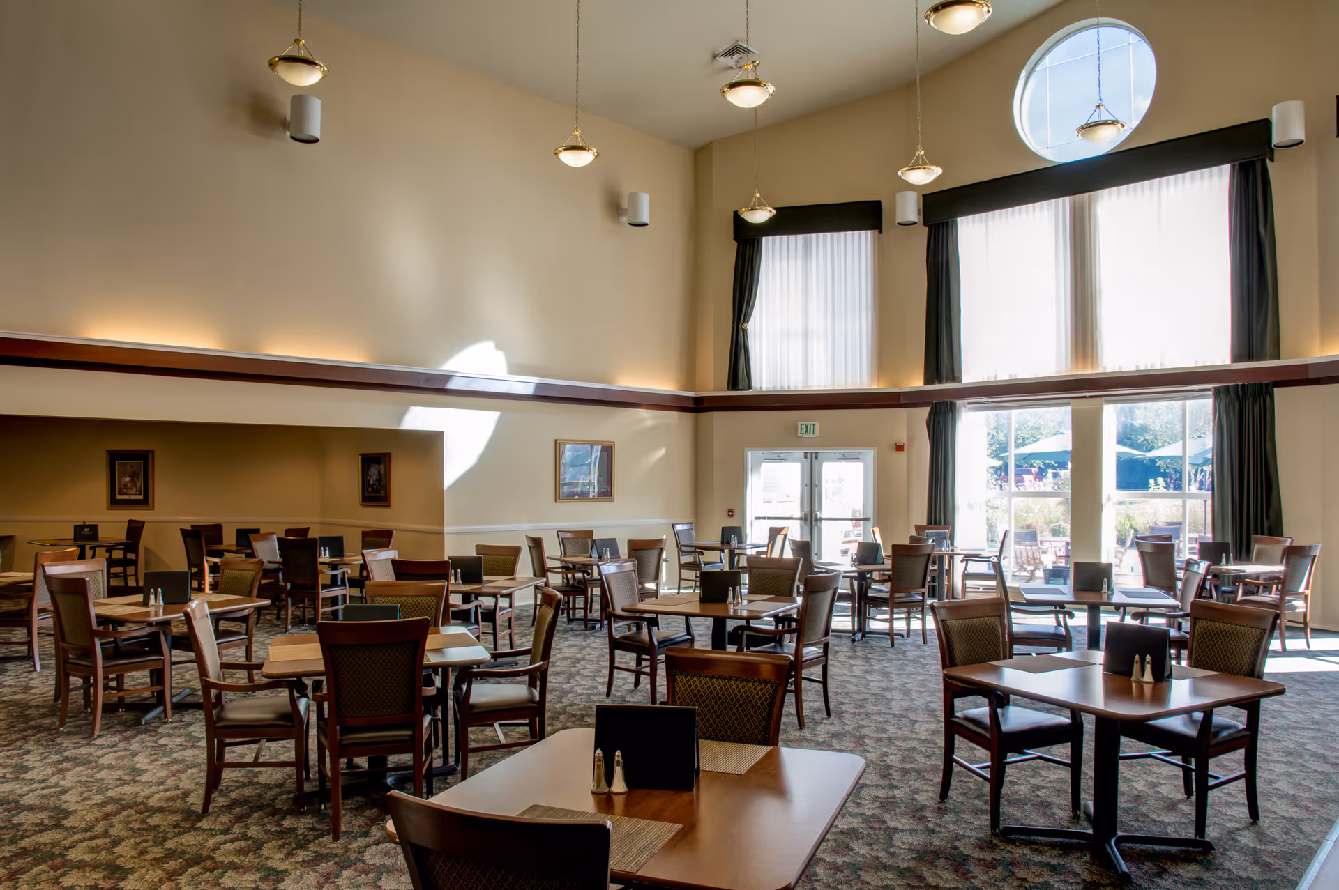 Spacious dining room with multiple tables and chairs, high ceilings, pendant lights, and large windows letting in natural light.