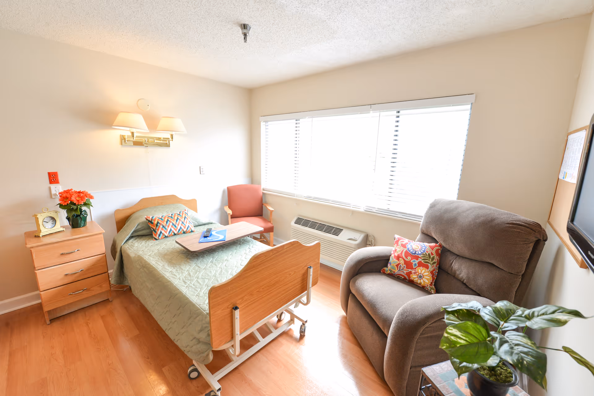 A small, bright room with a single hospital-style bed covered with a green quilt, a wooden nightstand with a clock and a vase of flowers, a red armchair, a large gray recliner with a colorful pillow, a window with blinds, and a small table with a potted plant.