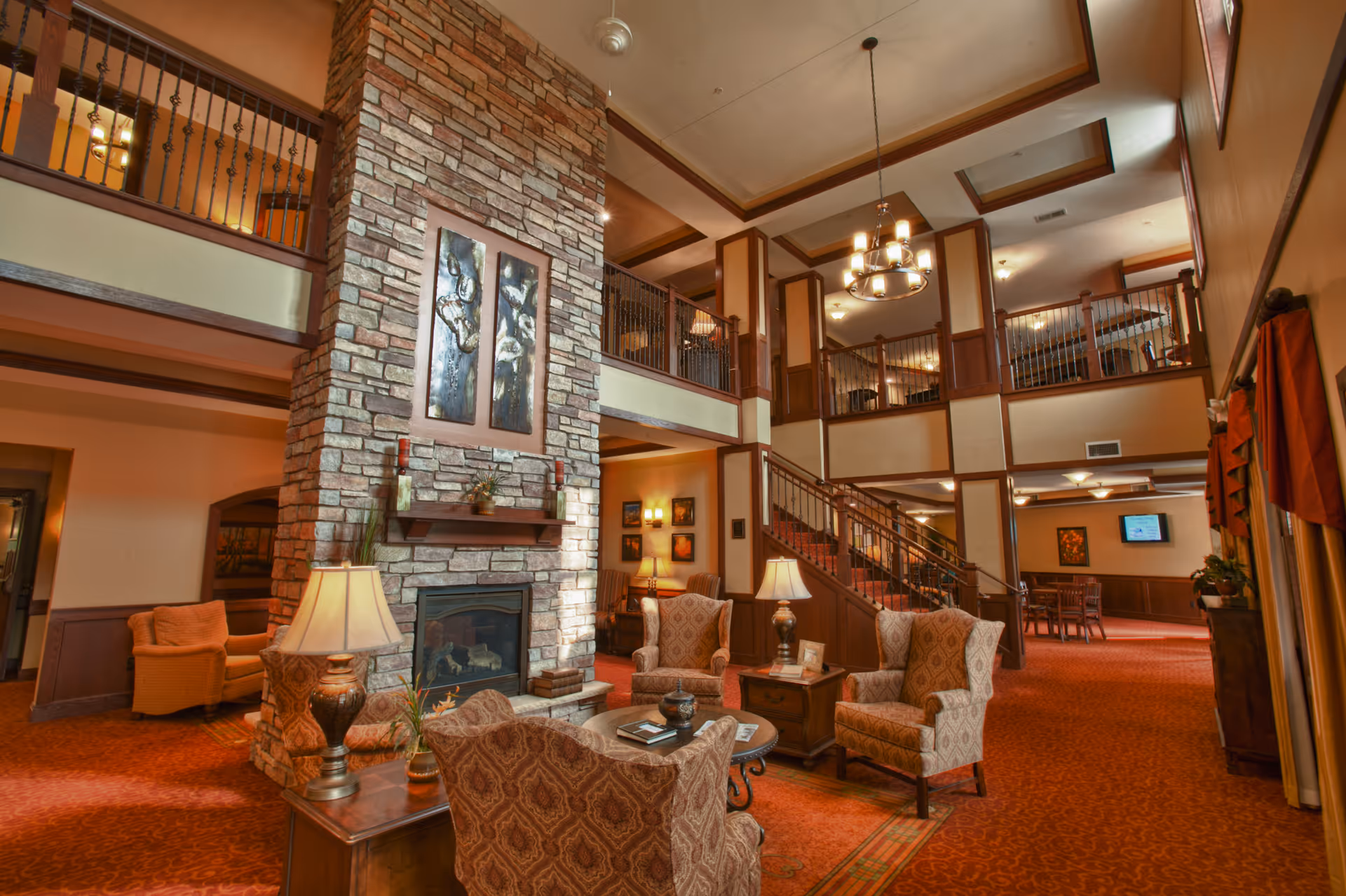 A spacious and warmly lit living room area with a tall stone fireplace in the center. The room features comfortable upholstered armchairs arranged around a coffee table, table lamps, and a staircase leading to an upper balcony. The decor includes framed artwork, warm earth tones, and a chandelier hanging from the ceiling.