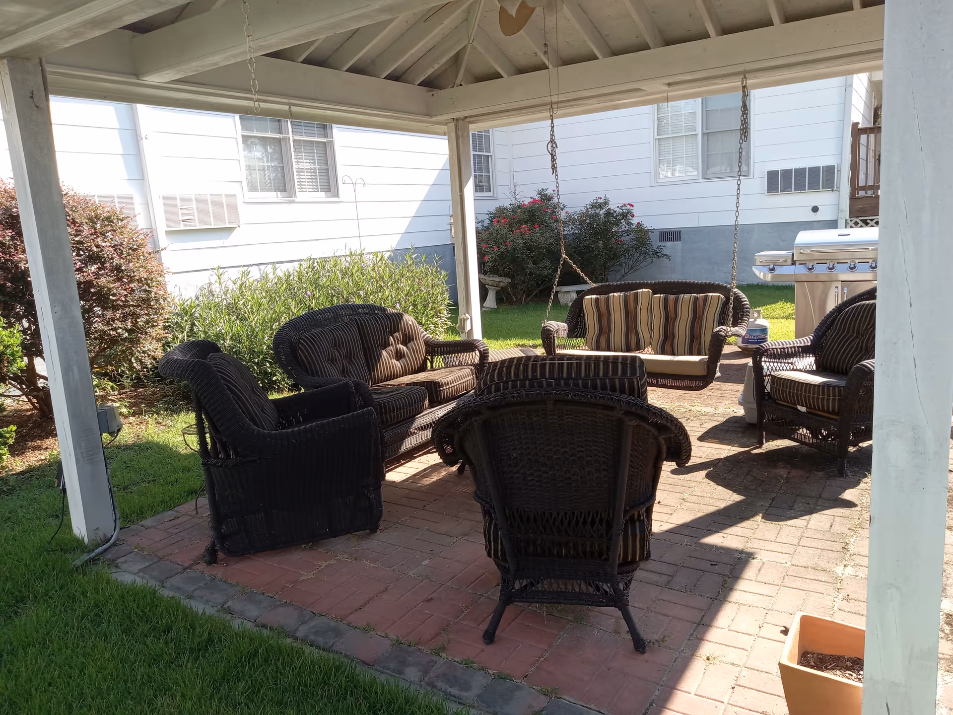 Covered outdoor patio with wicker chairs, a hanging couch swing, and a grill beside a white building.