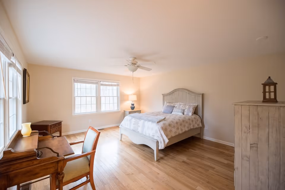 A bright bedroom with light wooden flooring, a large window letting in natural light, a white wooden bed with patterned bedding and pillows, a bedside table with a lamp, a wooden desk with a chair, and a white wooden dresser with a decorative lantern on top.