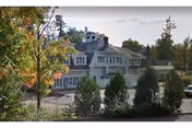 Exterior view of a two-story white nursing facility with dormer windows, a driveway, and trees in front.