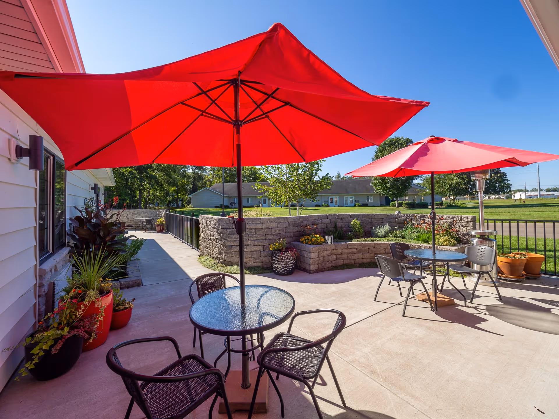 Outdoor patio area at Birch Meadows Senior Care with two round glass tables, each shaded by large red umbrellas. Black metal chairs surround the tables. There are potted plants along the building wall and a low stone wall with flowers and greenery in the background. The sky is clear and blue.