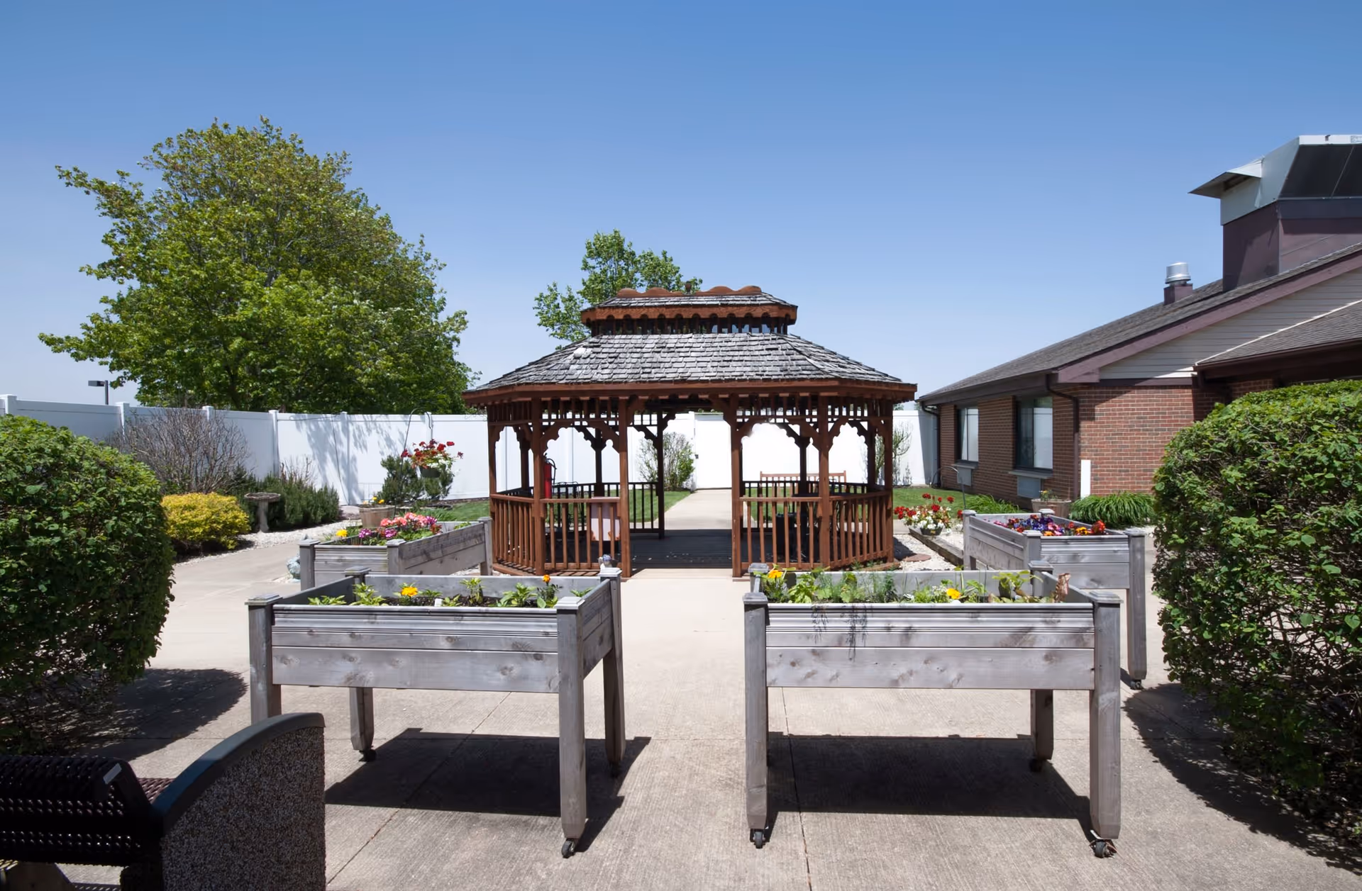 Outdoor garden area with a wooden gazebo in the center, surrounded by raised garden beds with plants and flowers. There are bushes and trees around the paved walkway, and a brick building is visible on the right side under a clear blue sky.