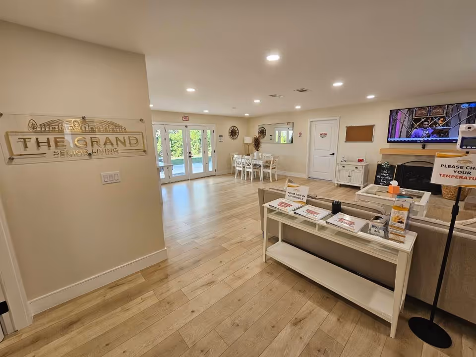Interior view of a senior living facility common area with light wood flooring, a seating area with a couch and table, a dining table with chairs near glass doors, a wall-mounted TV, and a sign on the wall that reads 'THE GRAND SENIOR LIVING'.