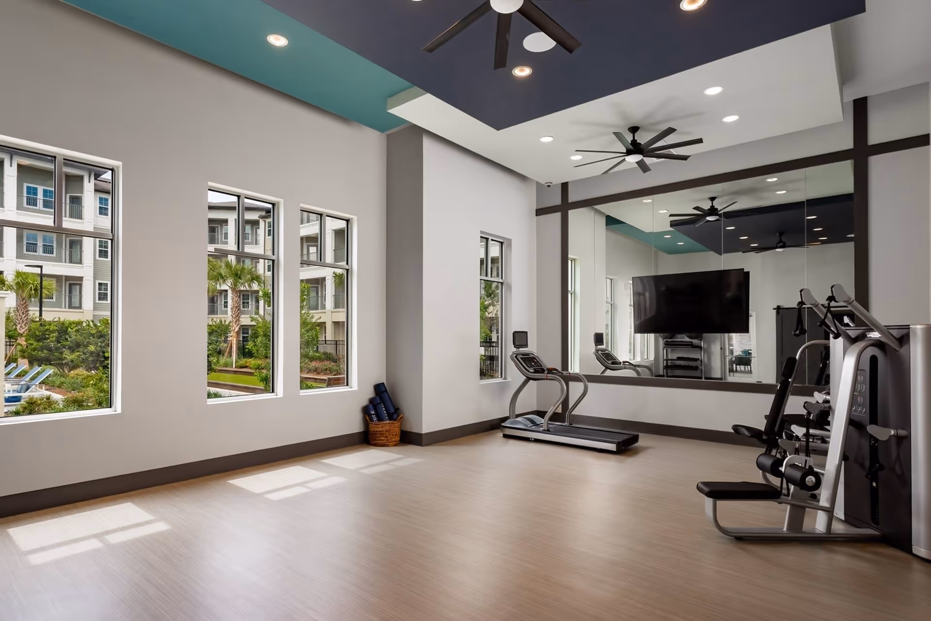 Bright fitness room with a treadmill, weight machine, large wall mirror and TV, ceiling fans, and windows overlooking an outdoor courtyard.
