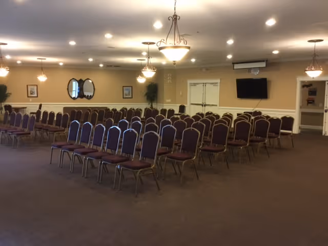 A large meeting room with rows of padded chairs arranged theater-style, chandeliers, a wall-mounted TV, and double doors.