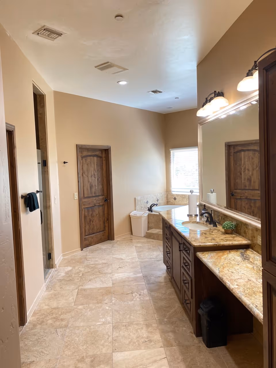 A spacious bathroom with beige tiled floor and walls painted in a light tan color. The room features a large granite countertop with two sinks, dark wood cabinetry, and a large mirror with three light fixtures above it. There is a corner bathtub with a window above it, a wooden door, and a towel rack with a dark towel hanging on it. The ceiling has recessed lighting and ventilation vents.