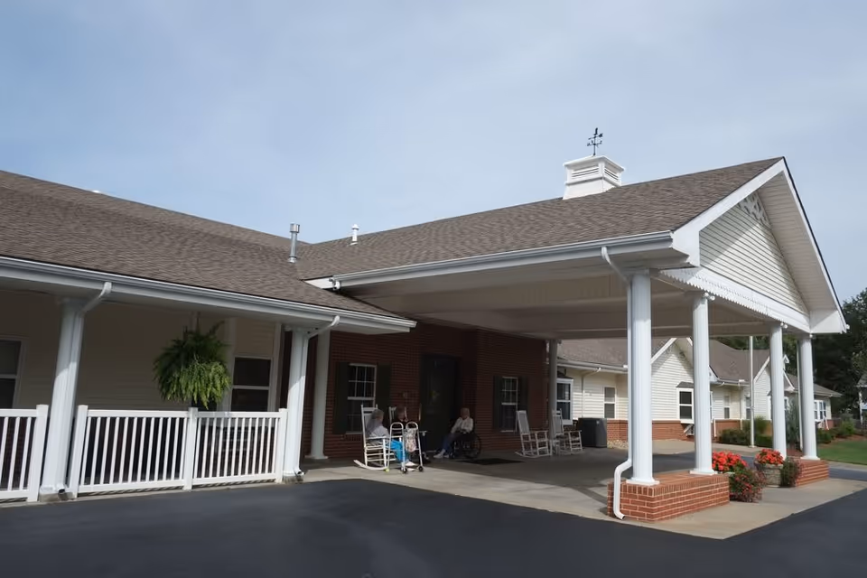 Covered front entrance of a single-story senior living facility with white columns, a driveway, and residents sitting on rocking chairs.