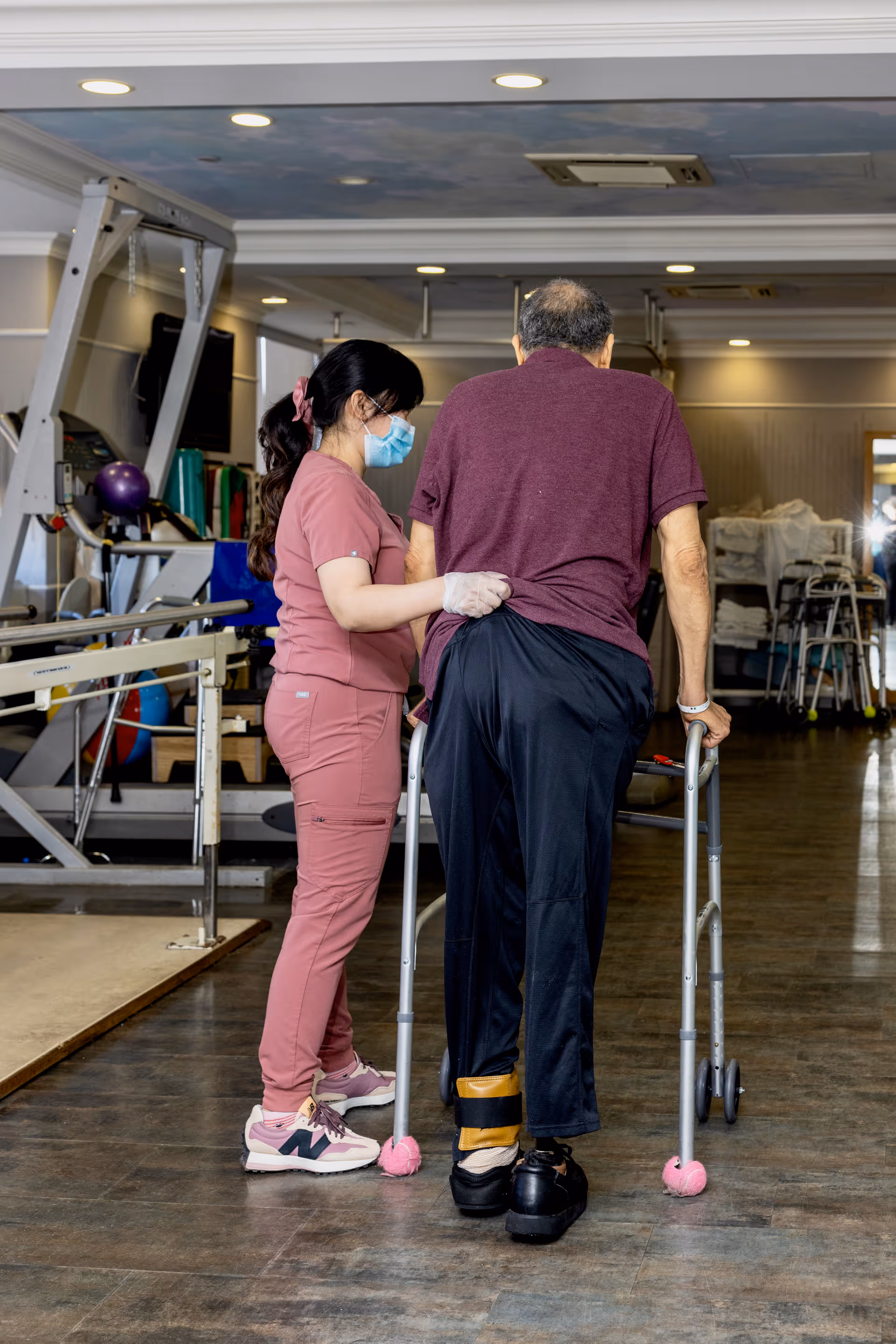 A healthcare worker wearing pink scrubs, a face mask, and gloves assists an elderly man using a walker in a rehabilitation or physical therapy room with exercise equipment in the background.