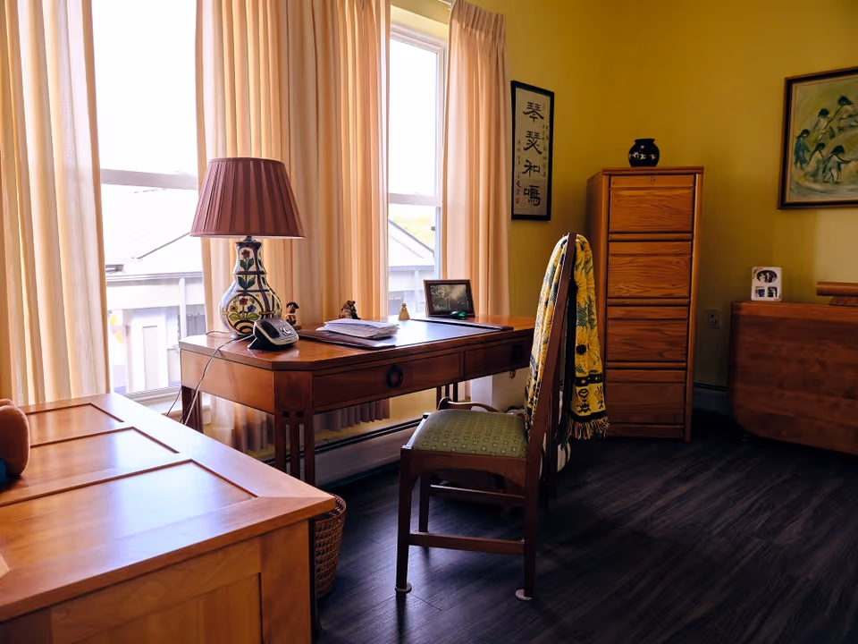 Sunlit room with a wooden desk and chair, lamp, dresser and curtained windows.