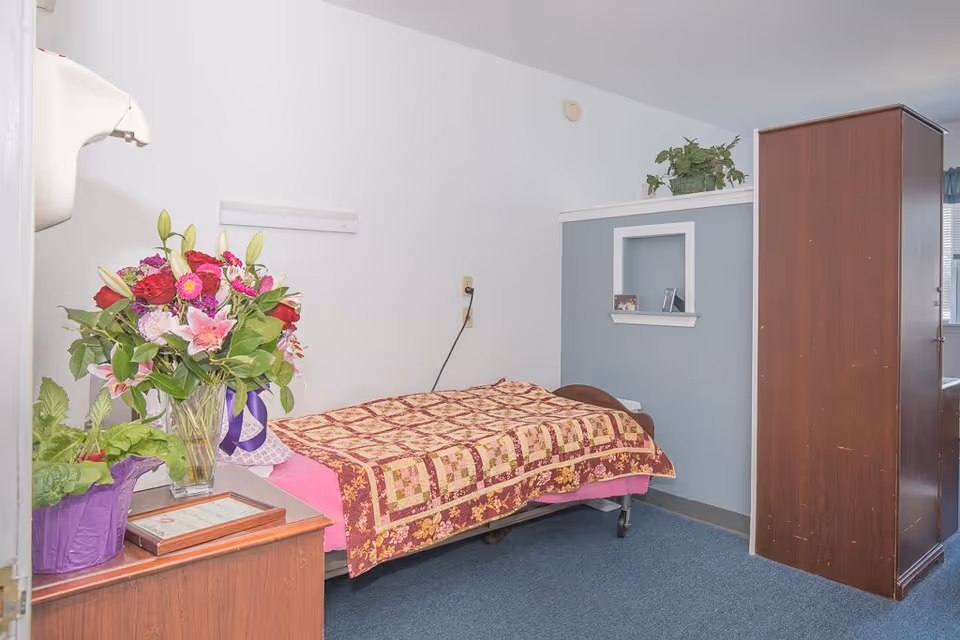 A small bedroom in a senior living facility with a single bed covered by a patterned quilt. There is a wooden cabinet on the right side and a small wooden table on the left with a large bouquet of flowers and a potted plant. The walls are white and blue, and there is a small recessed shelf with framed photos. The floor is carpeted in blue.