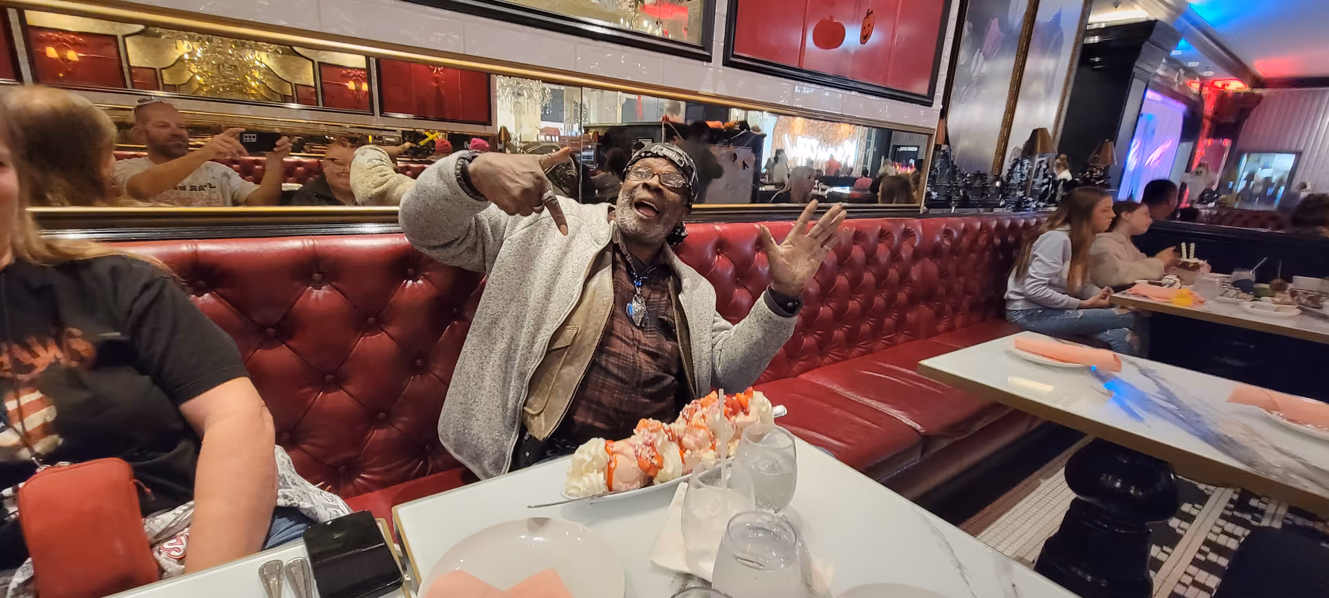A lively restaurant scene with a man sitting on a red tufted bench, smiling and gesturing enthusiastically. The table in front of him has plates, glasses of water, and a dessert with whipped cream and strawberries. Other diners are seated at tables in the background, and the walls are decorated with mirrors and framed pictures.