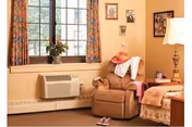 A cozy living area with a large window featuring patterned curtains, a brown recliner chair with a pink hat and white cloth draped over it, a small side table with a lamp and decorative items, and a bed partially visible with a peach-colored blanket. There is also a potted plant on the window sill and a pair of slippers on the floor.