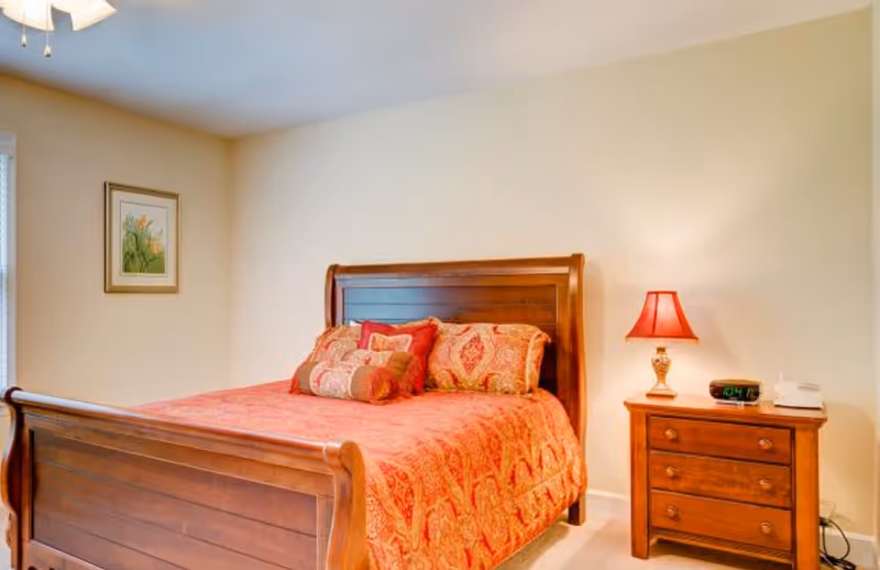 A bedroom with a wooden sleigh bed covered in a red and orange patterned bedspread and multiple decorative pillows. Next to the bed is a wooden nightstand with a red lampshade, a digital alarm clock, and a telephone. A framed floral picture hangs on the wall, and a ceiling fan is partially visible.