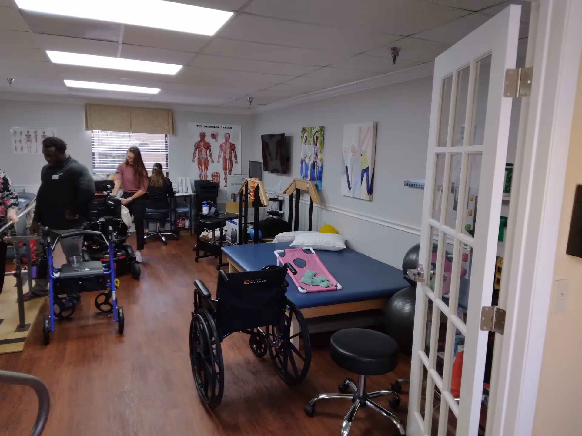 A physical therapy or rehabilitation room with exercise equipment, a wheelchair, a walker, a therapy bed with pillows, and posters of the muscular system and elderly people exercising on the walls. Two people are present, one assisting another using a walker.