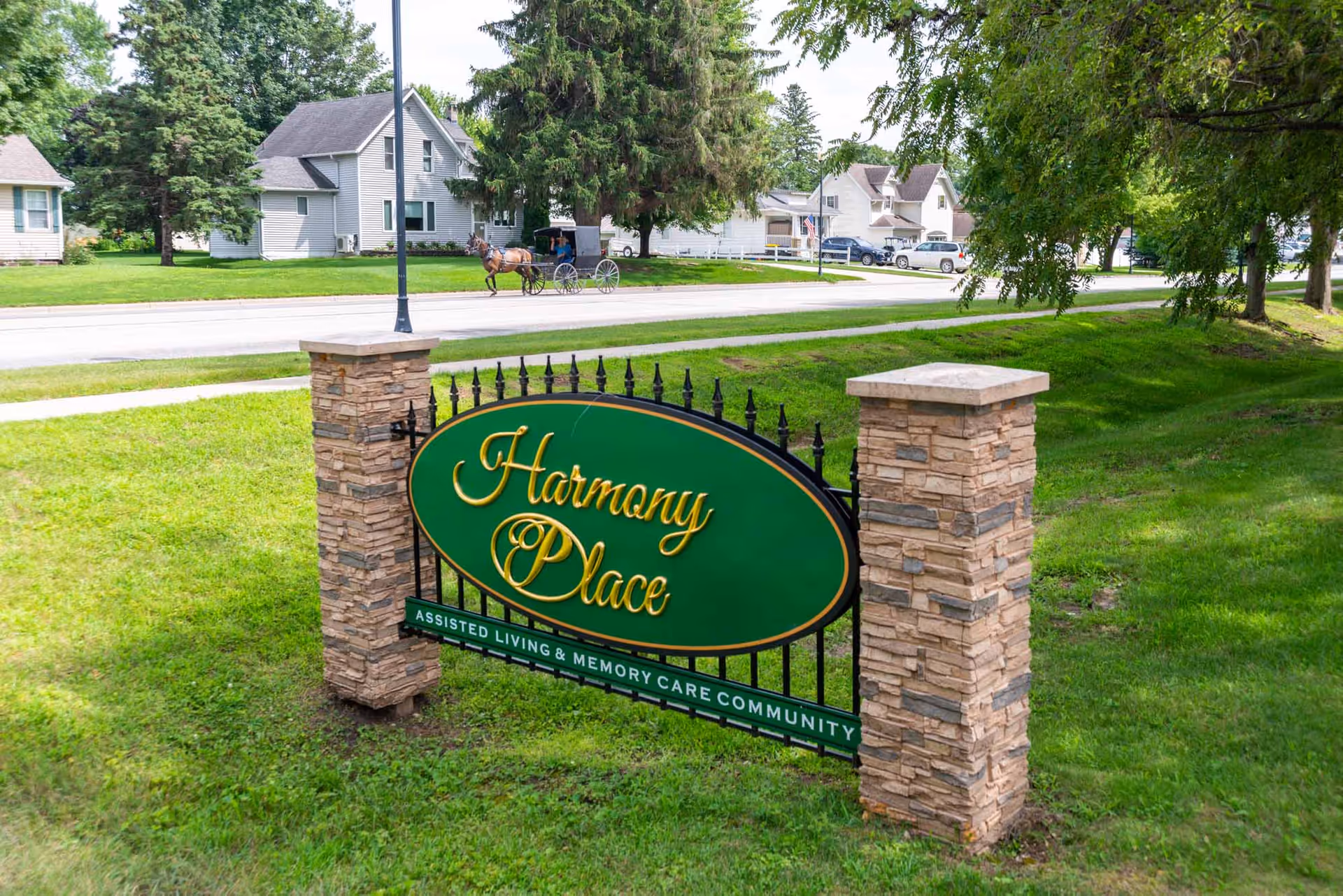Green oval sign reading "Harmony Place" mounted between stone pillars on a grassy lawn with houses and a horse-drawn buggy in the background.