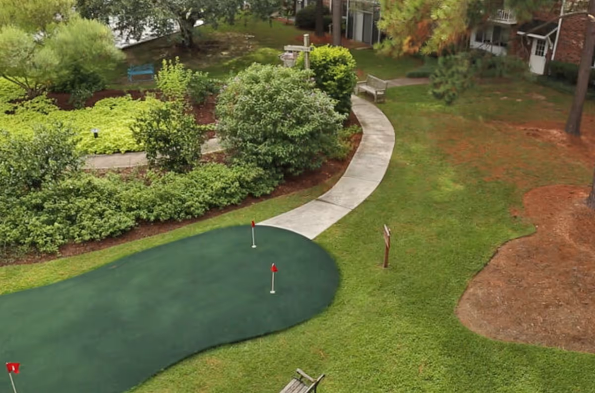 A well-maintained outdoor garden area with a putting green featuring two red flags, surrounded by green grass, bushes, and trees. There is a curved concrete pathway with benches along the side, and a building with brick walls and white doors is visible in the background.