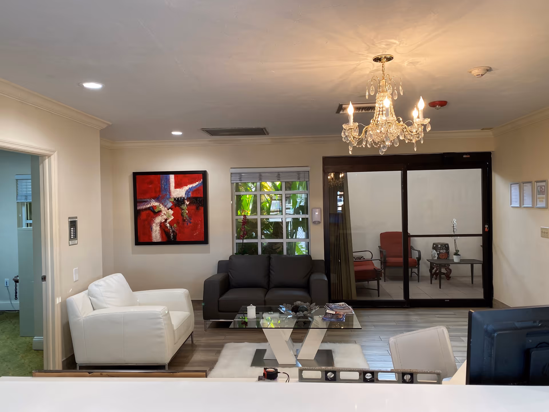 A reception lounge with white and gray sofas, a glass coffee table, a chandelier, colorful wall art, and sliding glass doors to a patio seating area.
