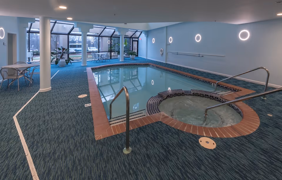 Carpeted indoor swimming pool with an attached circular hot tub in a room with columns, seating, and large windows.
