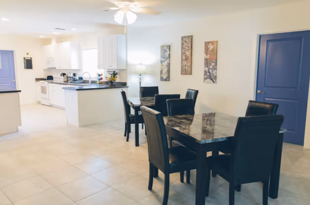 Interior view of a dining area and kitchen in a senior living facility. The dining area has two tables with black chairs, and the kitchen features white cabinets, a stove, and a countertop with a fruit bowl. The walls are decorated with three floral paintings, and there are two blue doors visible.