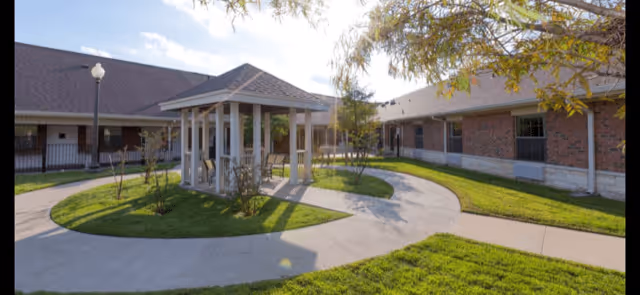Outdoor courtyard area at Legend Oaks Healthcare and Rehabilitation - Ennis featuring a paved walkway, green grass, a small gazebo with benches, and surrounding single-story brick buildings under a partly cloudy sky.