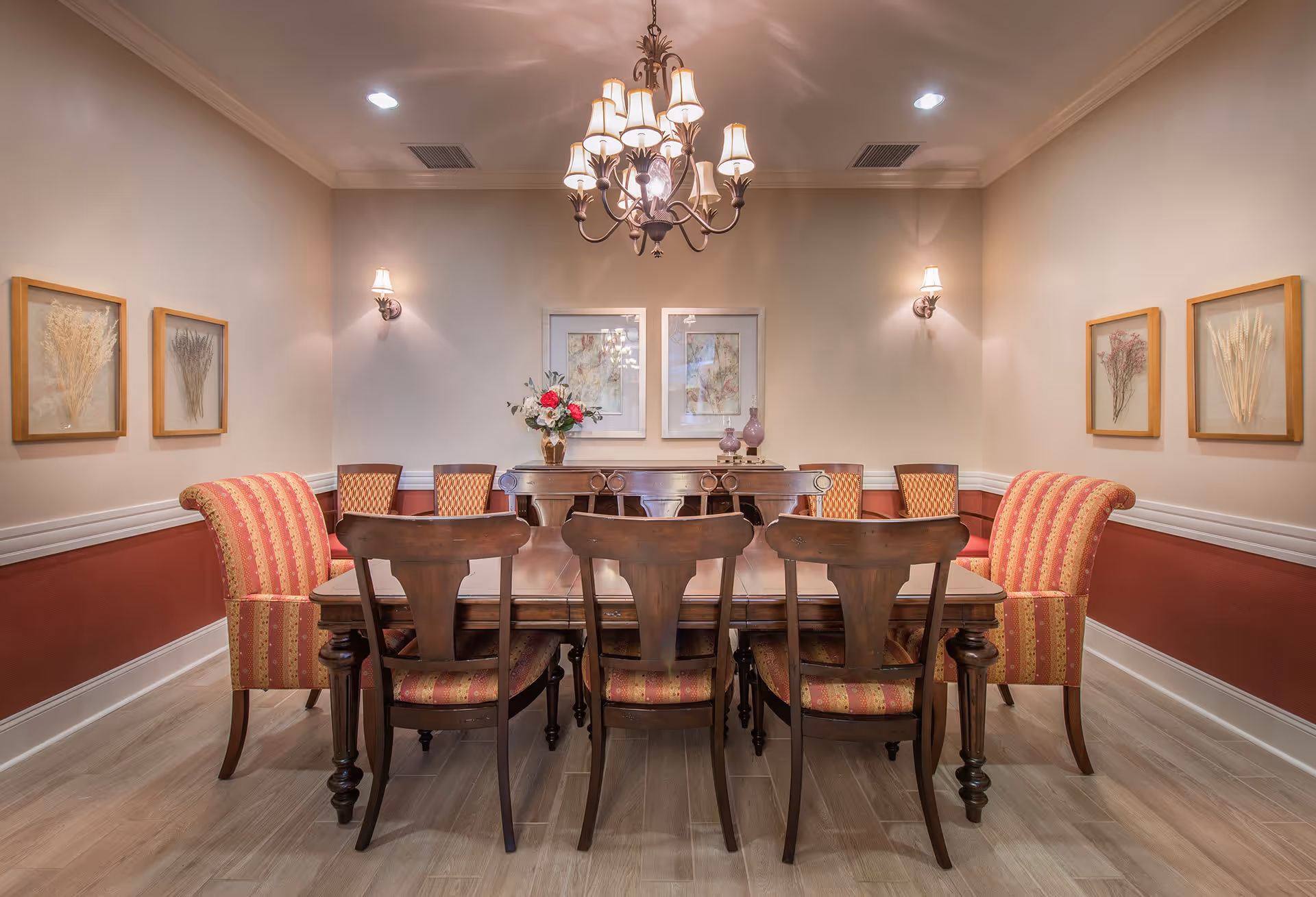 A formal dining room with a wooden dining table surrounded by six chairs, two of which have red and gold striped upholstery. The room features a chandelier hanging from the ceiling, two wall sconces, framed botanical artwork on the walls, and a sideboard with decorative vases and a flower arrangement.