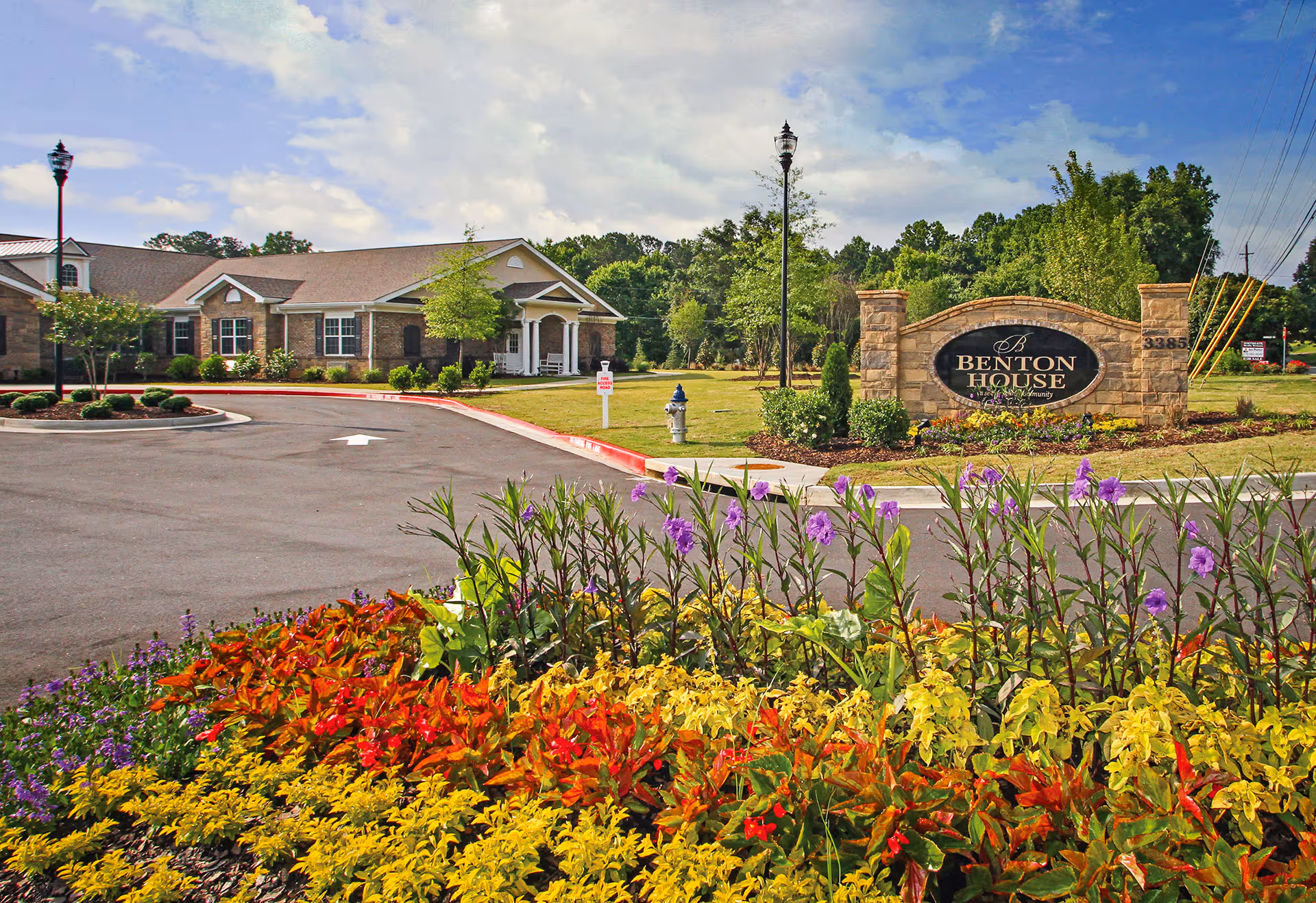 Entrance to Benton House senior living community with a stone sign, colorful flower bed in the foreground, a paved driveway, and a single-story brick building under a partly cloudy sky.