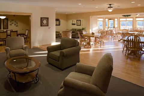 Well-lit communal room with upholstered armchairs in the foreground and multiple wooden dining tables near large windows.